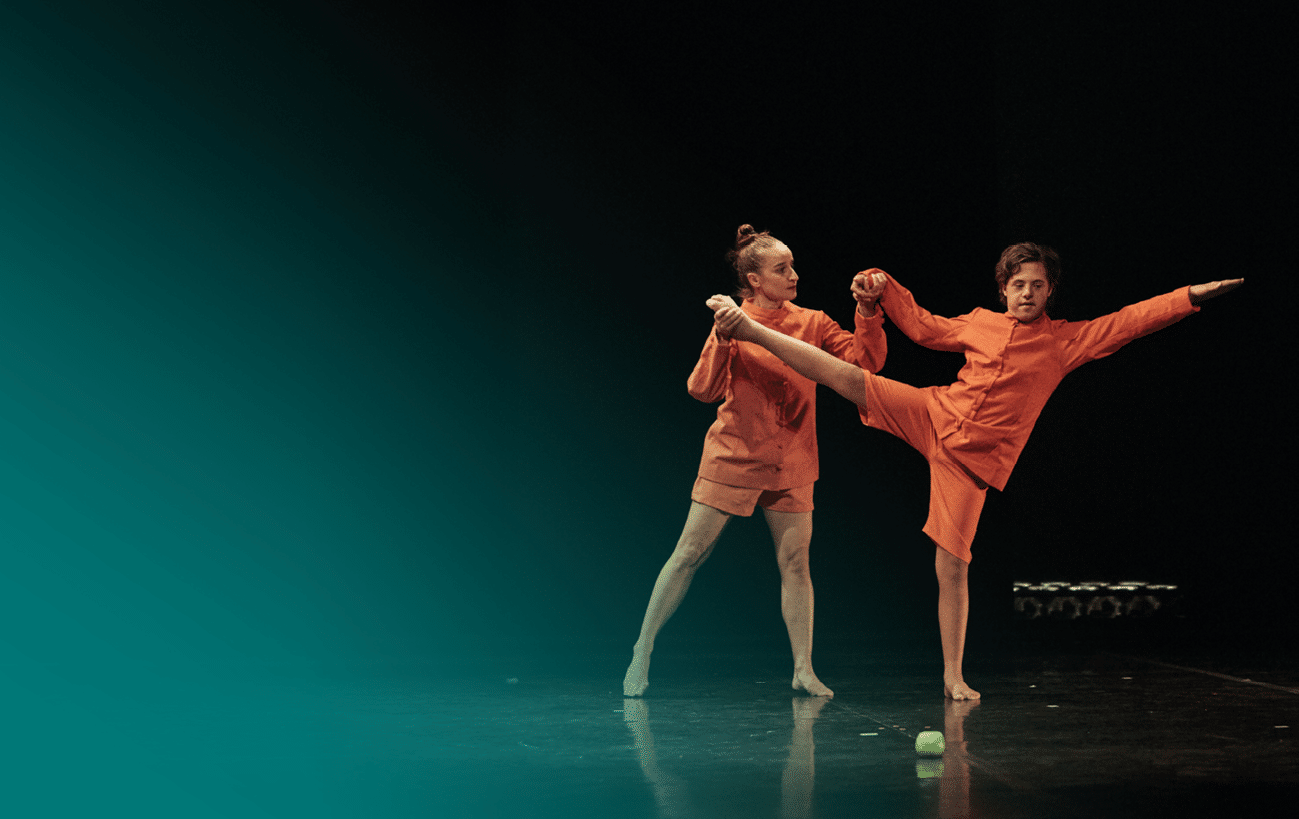 Dos personas vestidas con ropa naranja realizan una coreograf a de danza contempor nea en un escenario oscuro con un objeto verde en el suelo. Descripci n generada con IA
