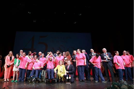 Un grupo grande de personas vestidas con ropa formal y camisetas rosas posan juntas en un escenario durante un evento conmemorativo. Descripci n generada con IA