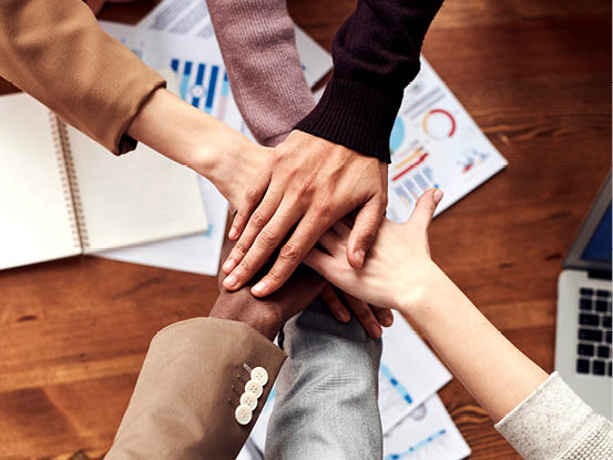 Cinco personas unen sus manos sobre una mesa con documentos y una computadora port til en un ambiente de trabajo colaborativo. Descripci n generada con IA