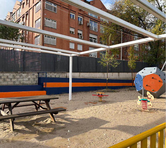Parque infantil vac o con suelo de arena, juegos de resorte, una estructura para escalar y bancos de madera junto a un edificio de ladrillo. Descripci n generada con IA